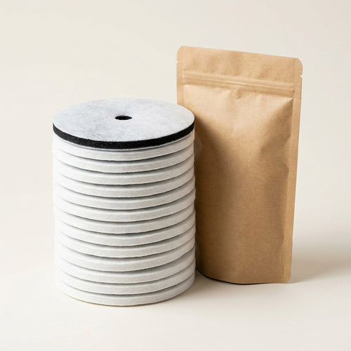 Stack of twelve round cat fountain replacement filters beside a kraft pouch, showing the white cotton face and charcoal carbon layer
